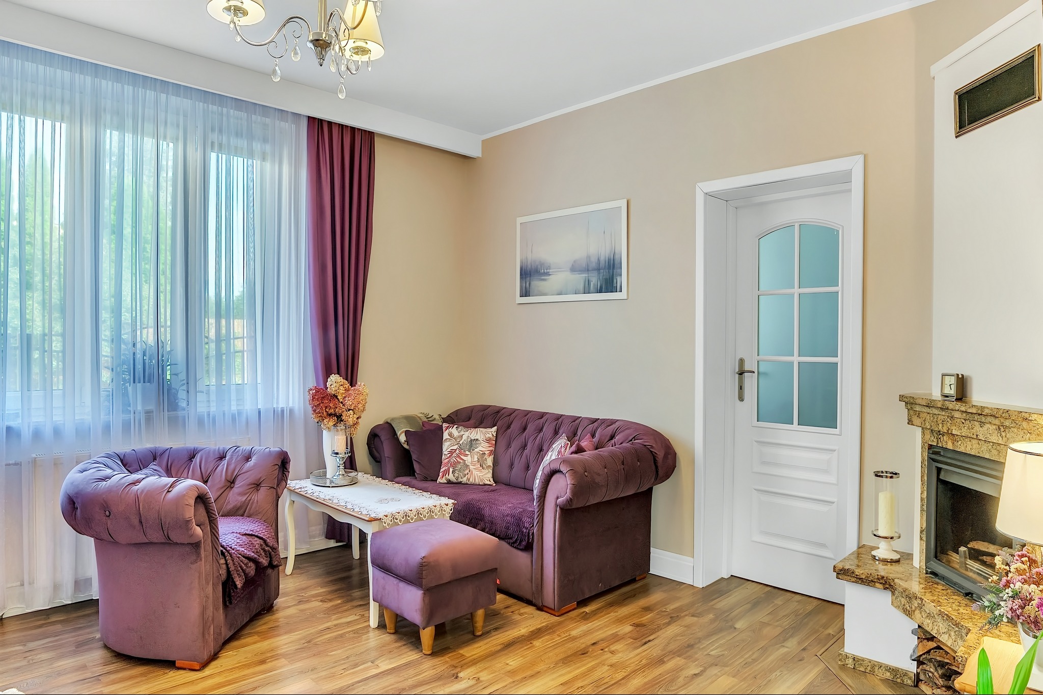 Salon w odcieniach fioletu i beżu: sofa Chesterfield, fotel z pufą, kominek z marmuru i obraz nad sofą.