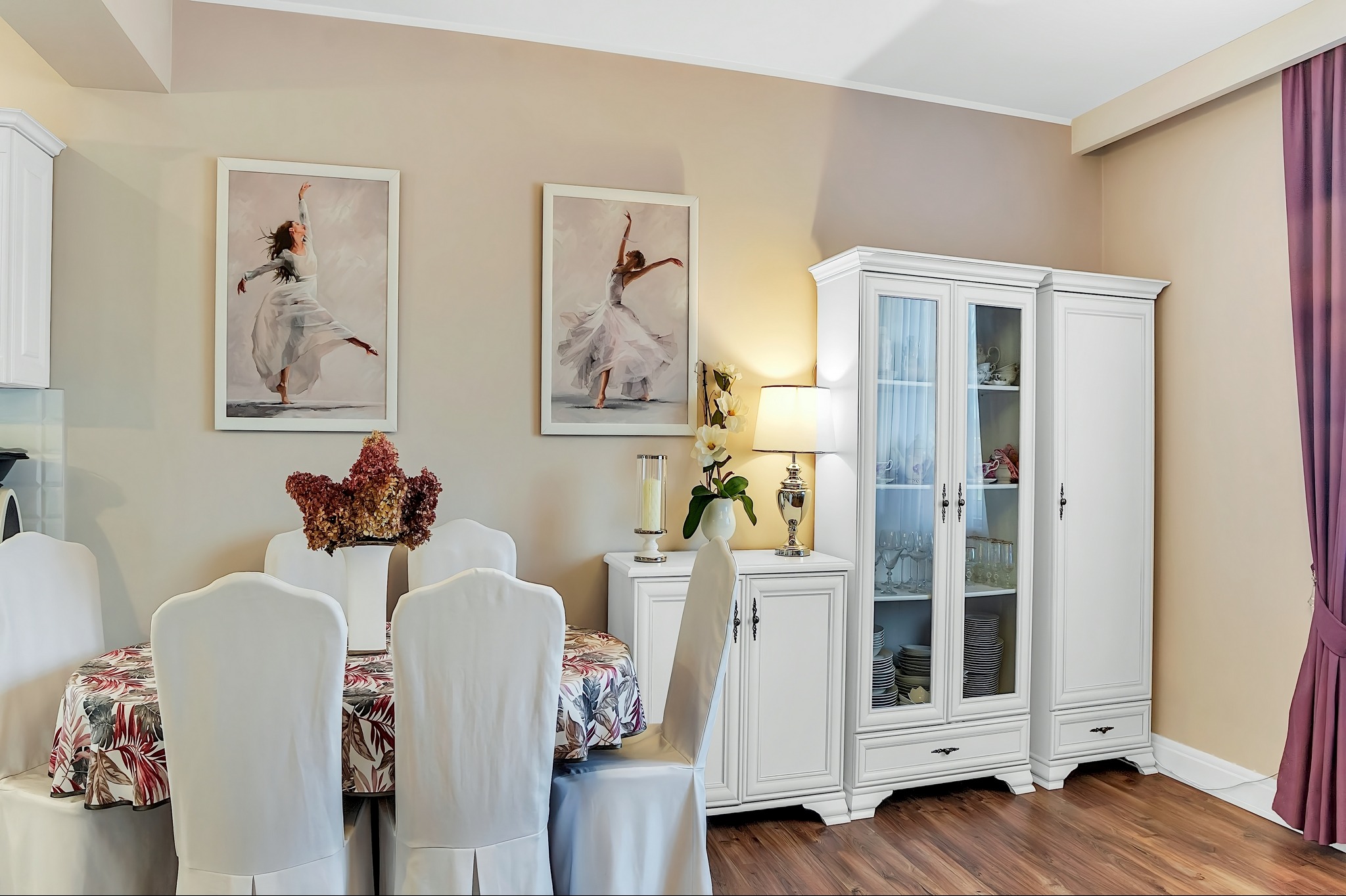 Elegancka jadalnia z białymi meblami, stołem nakrytym w kwiaty, obrazami baletnic i dekoracyjną lampą na komodzie.