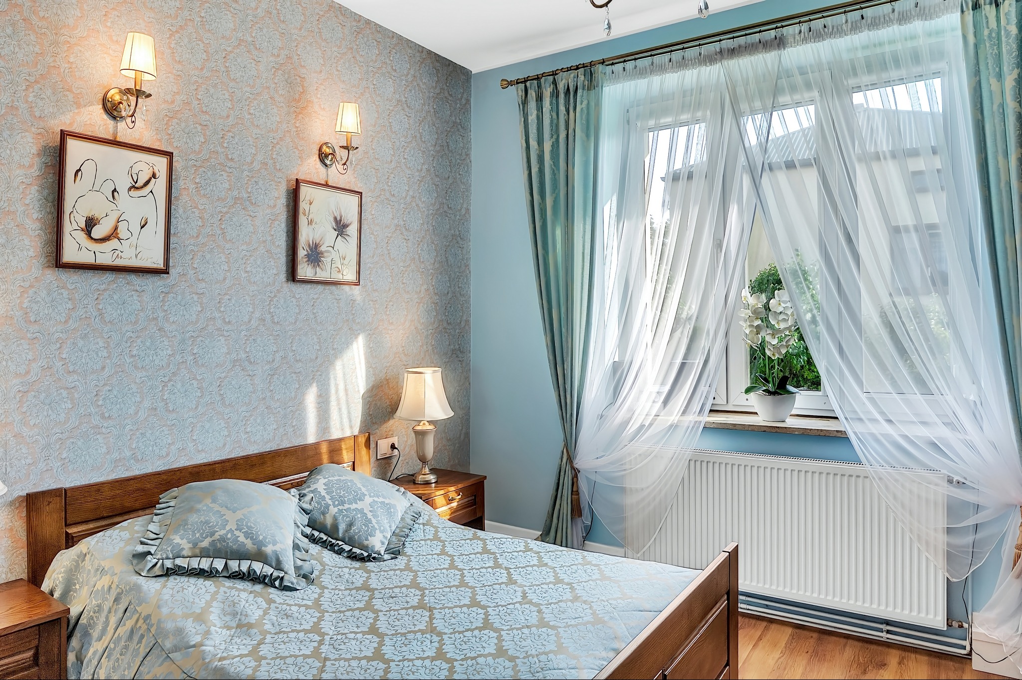 Przytulna sypialnia w błękitach: drewniane łóżko z wzorzystą pościelą, okno z firanami i zasłonami, lampy ścienne nad łóżkiem, kwiat na parapecie.