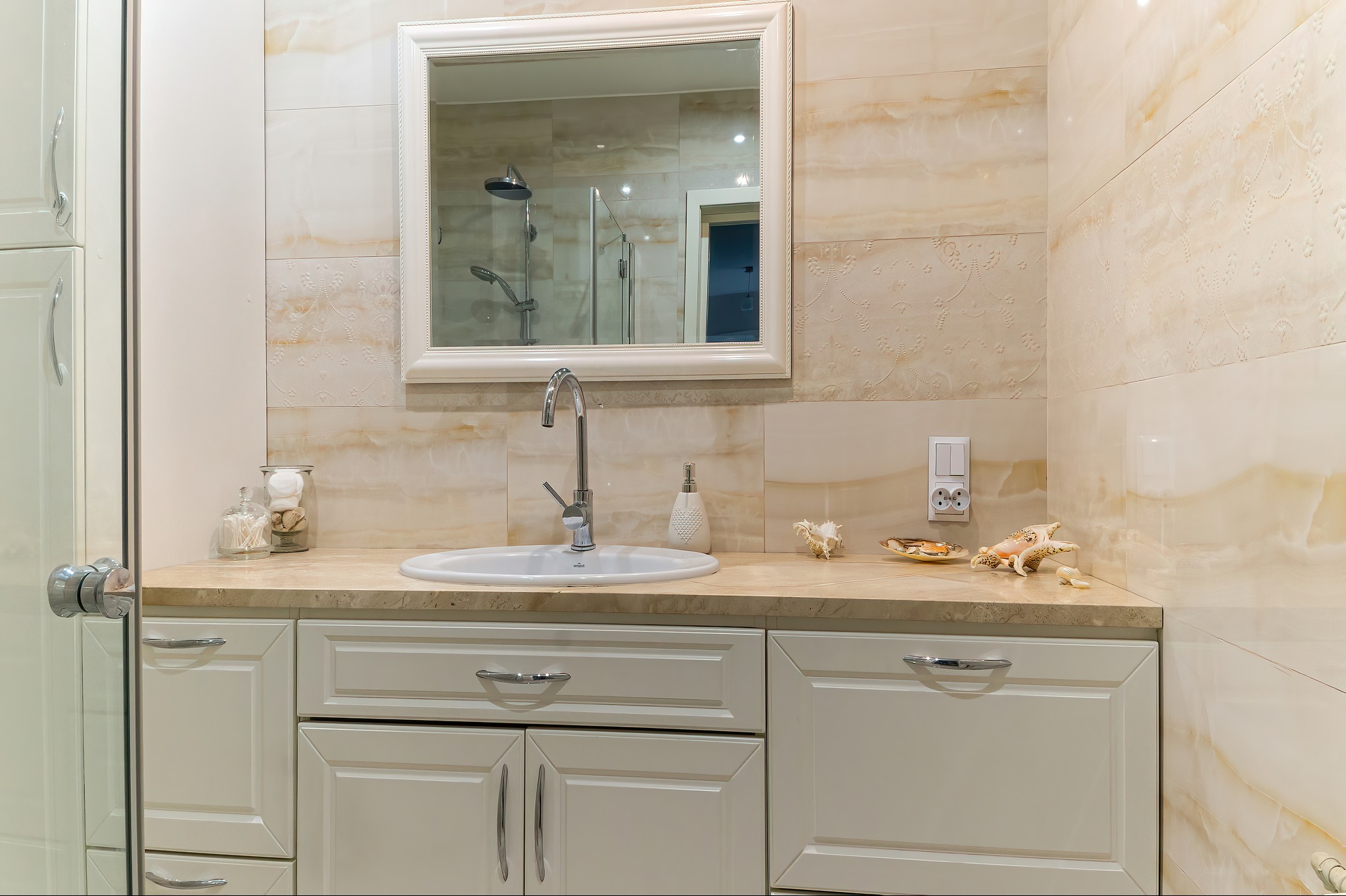 Jasna łazienka z umywalką wpuszczoną w blat, lustro w białej ramie odbija kabinę prysznicową, dekoracyjne muszle na blacie, płytki imitujące marmur z delikatnym wzorem.