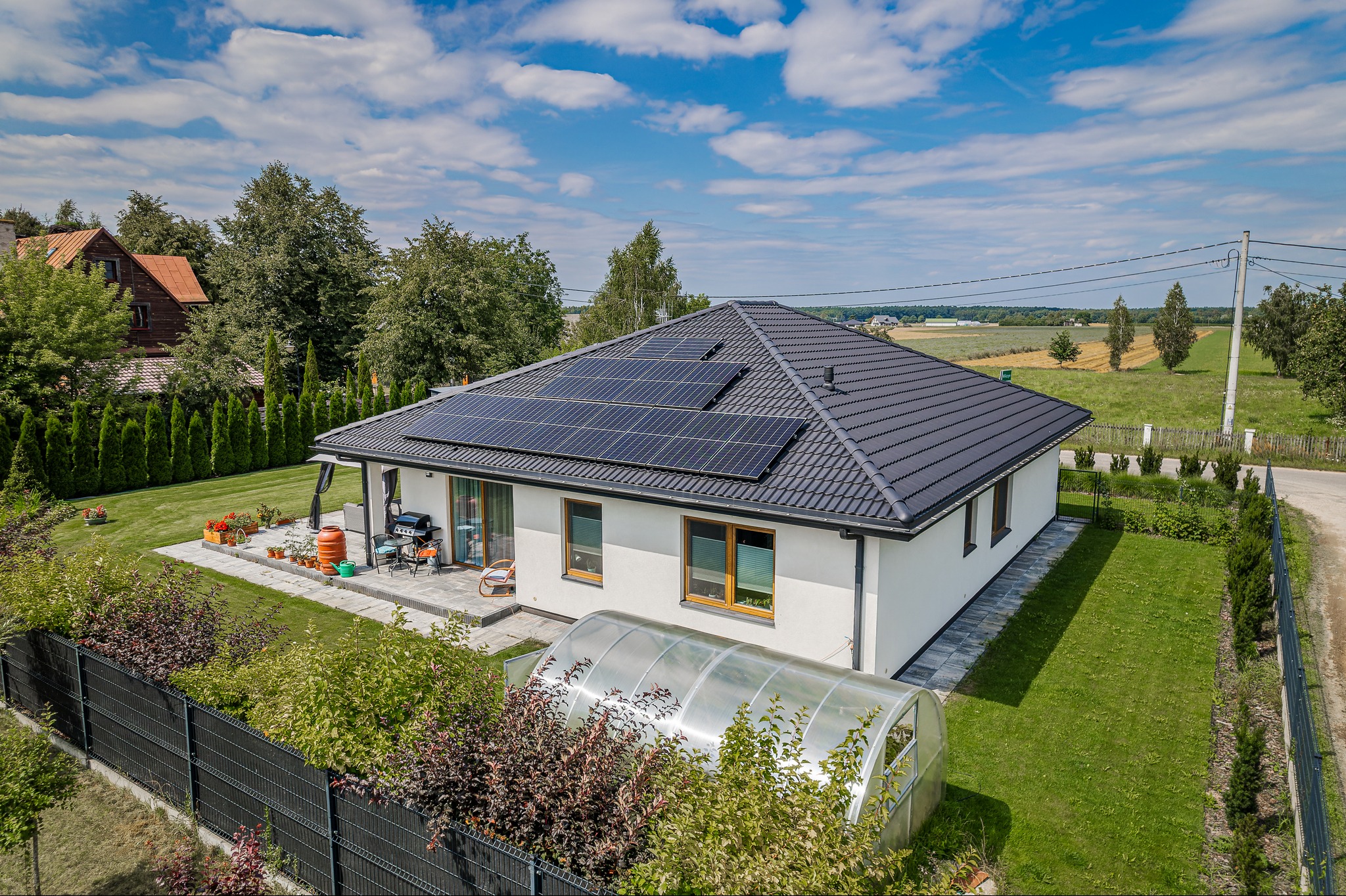 Nowoczesny dom jednorodzinny z panelami słonecznymi na dachu, widok z lotu ptaka, ogród z tunelem foliowym i zadbanym trawnikiem.
