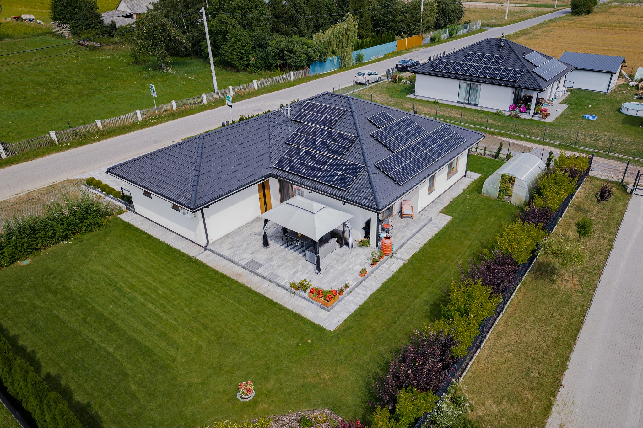 Widok z drona na nowoczesny dom z panelami słonecznymi na dachu, zadbanym trawnikiem, altaną ogrodową i szklarnią w tle.
