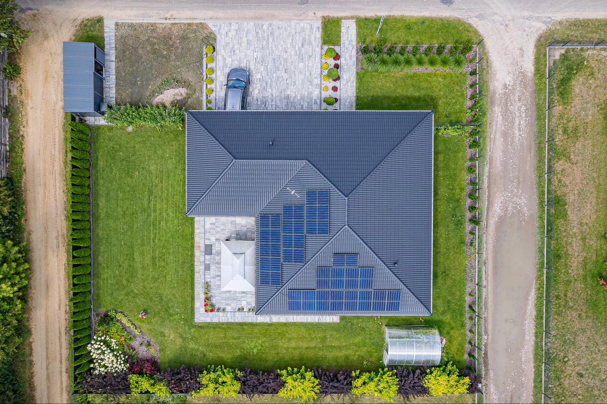 Zdjęcie z drona przedstawia widok z góry na dom z ciemnoszarą dachówką, na której zamontowane są panele słoneczne, zadbany trawnik, szary podjazd z kostki brukowej, szklarnię i samochód osobowy.