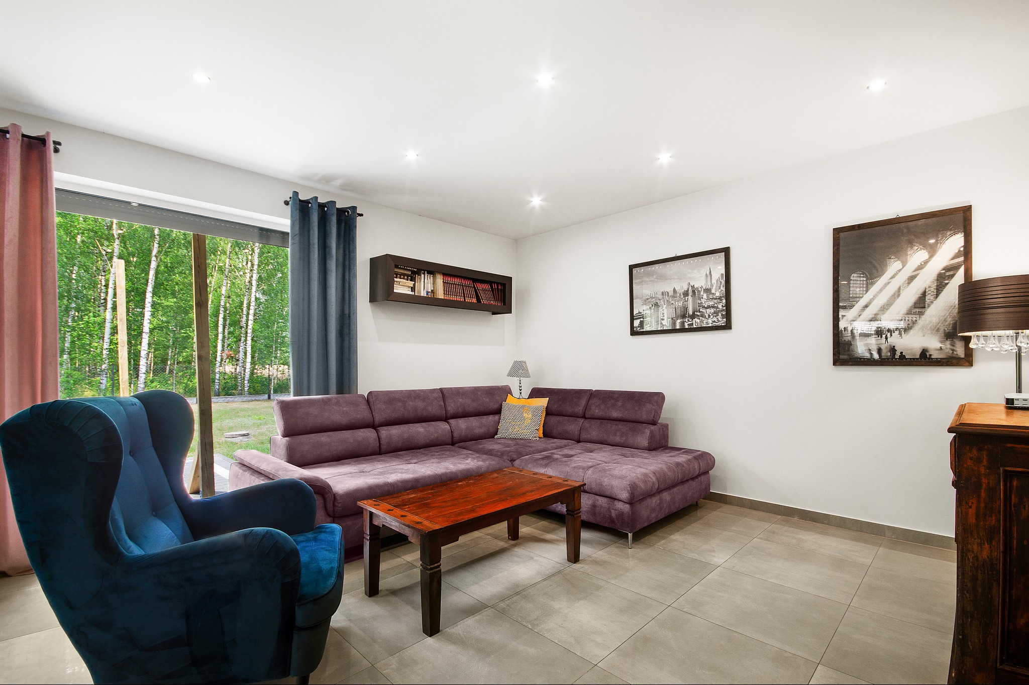 Jasny salon z narożną, fioletową kanapą, drewnianym stolikiem kawowym, niebieskim fotelem typu uszak i widokiem na las za oknem.