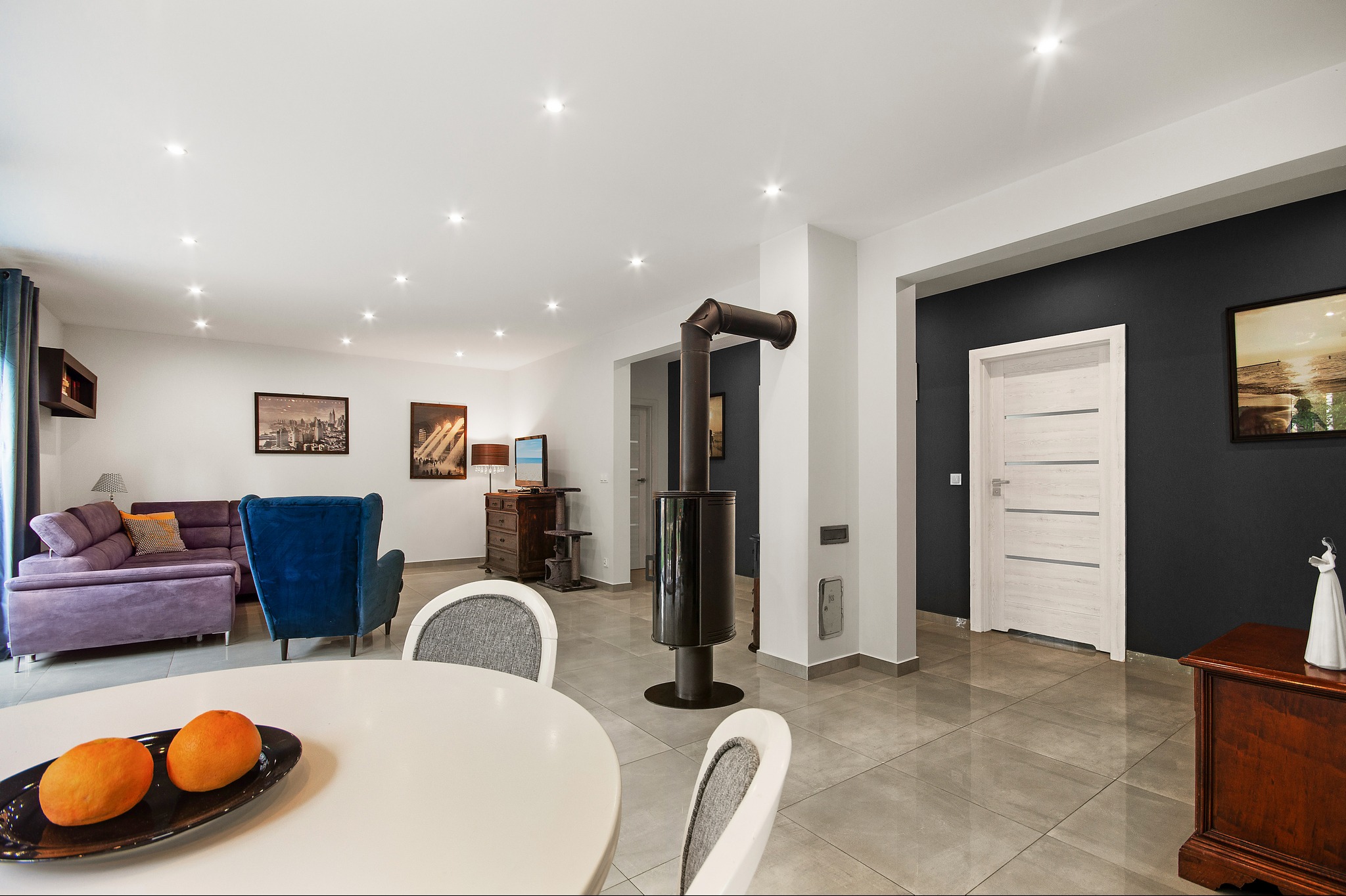 Przestronny salon z nowoczesnym piecem wolnostojącym, fioletową sofą, niebieskim fotelem i białym stołem z owocami na pierwszym planie.