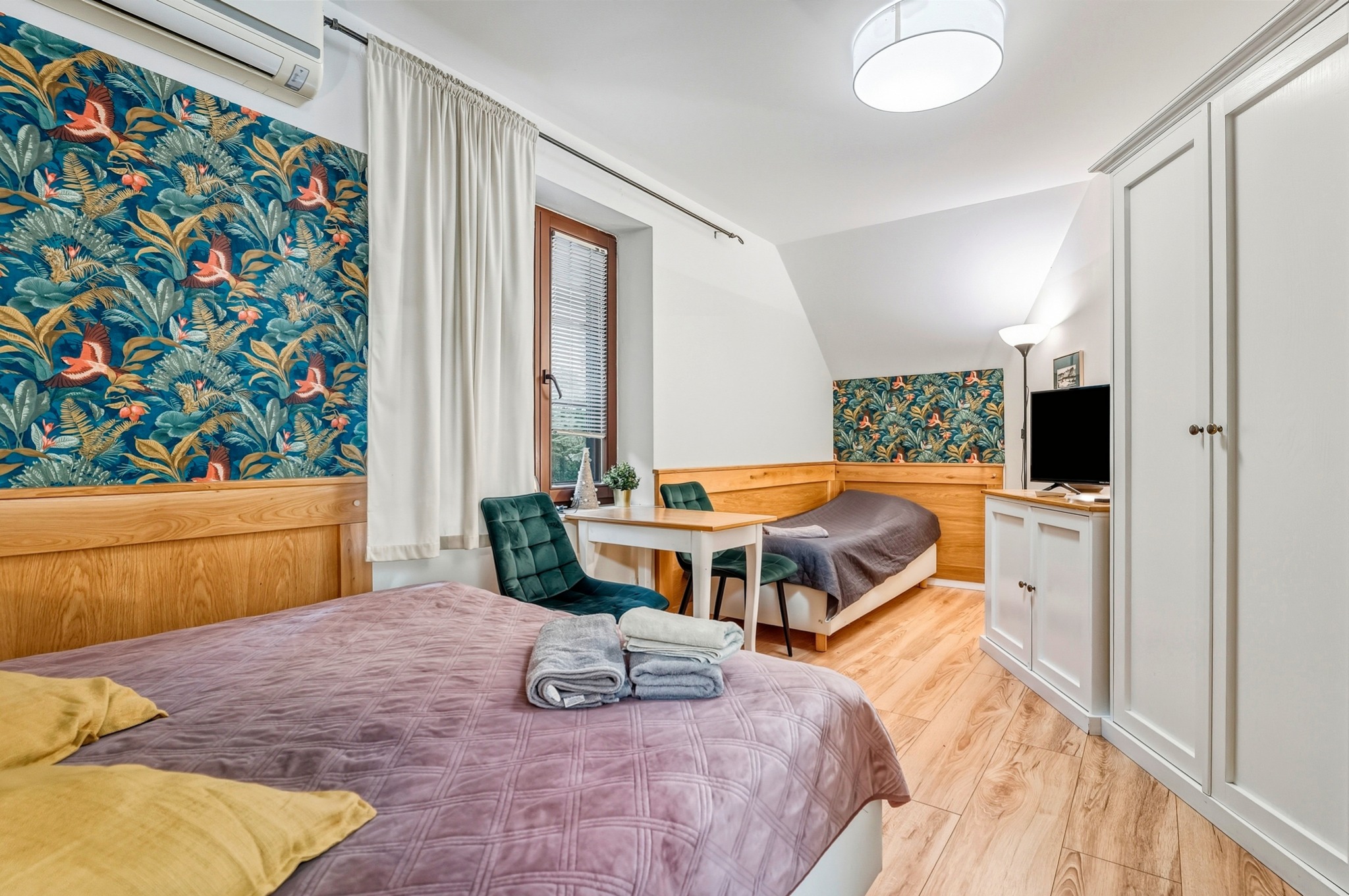 Przytulny pokój gościnny z dwoma łóżkami, drewnianą podłogą i szafą w zabudowie. Ściany zdobi tapeta w tropikalne wzory. Widoczne detale: ręczniki, stolik z krzesłami.
