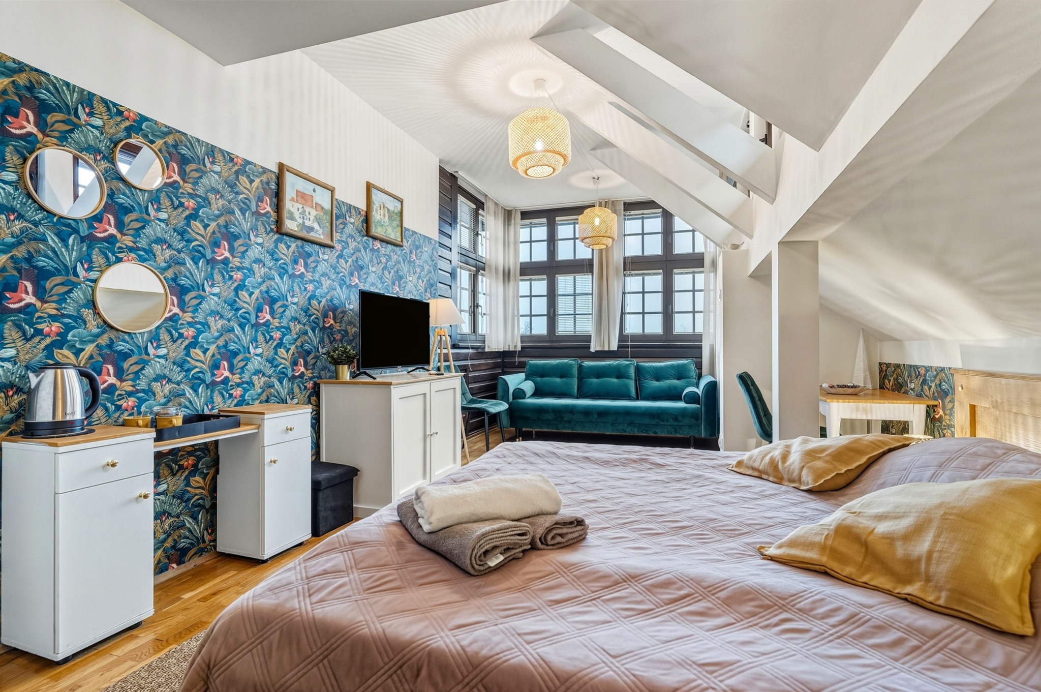 Sypialnia na poddaszu z wzorzystą tapetą, okrągłymi lustrami i turkusową sofą. Widok z łóżka na okno i lampy. Nowoczesny design z elementami retro.