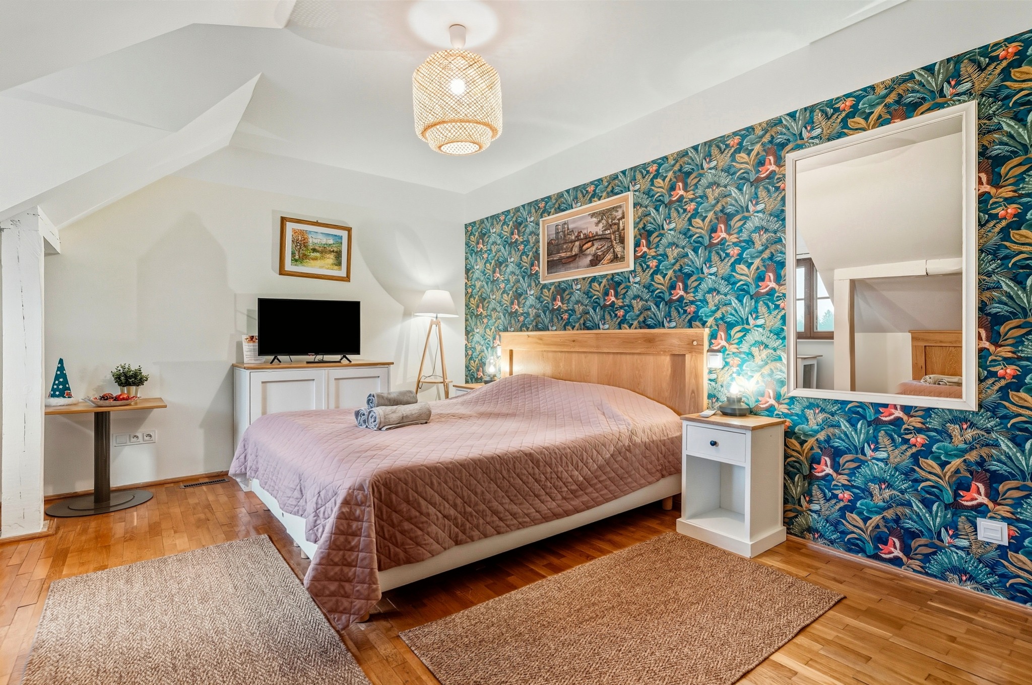 Sypialnia na poddaszu z łóżkiem, wzorzystą tapetą w tropikalne motywy i drewnianą podłogą. Widoczne lampki nocne i dywaniki.