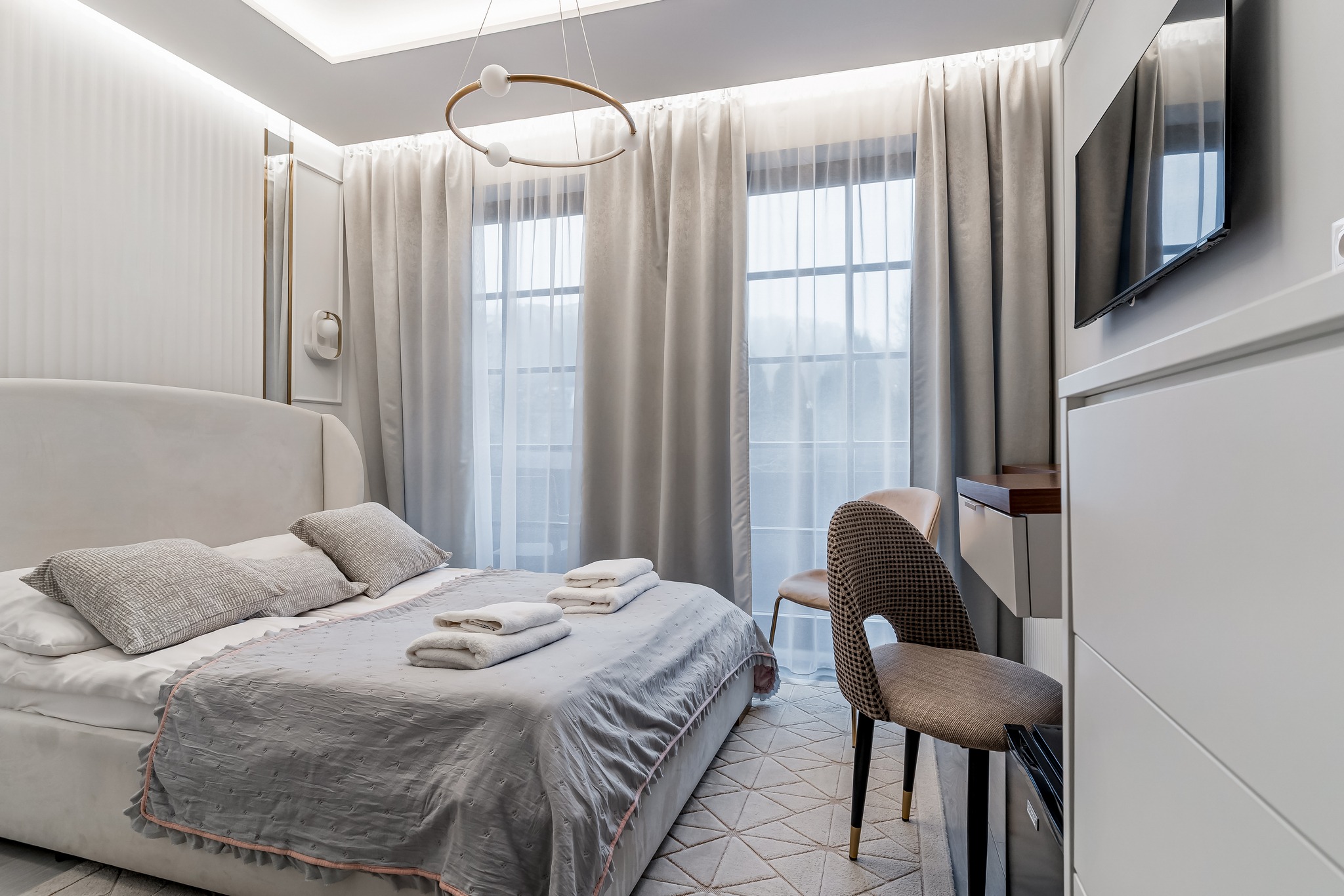 Jasna sypialnia w neutralnych barwach: łóżko z szarą narzutą, okno z zasłonami, krzesło i biurko. Minimalistyczny design, nowoczesne oświetlenie.