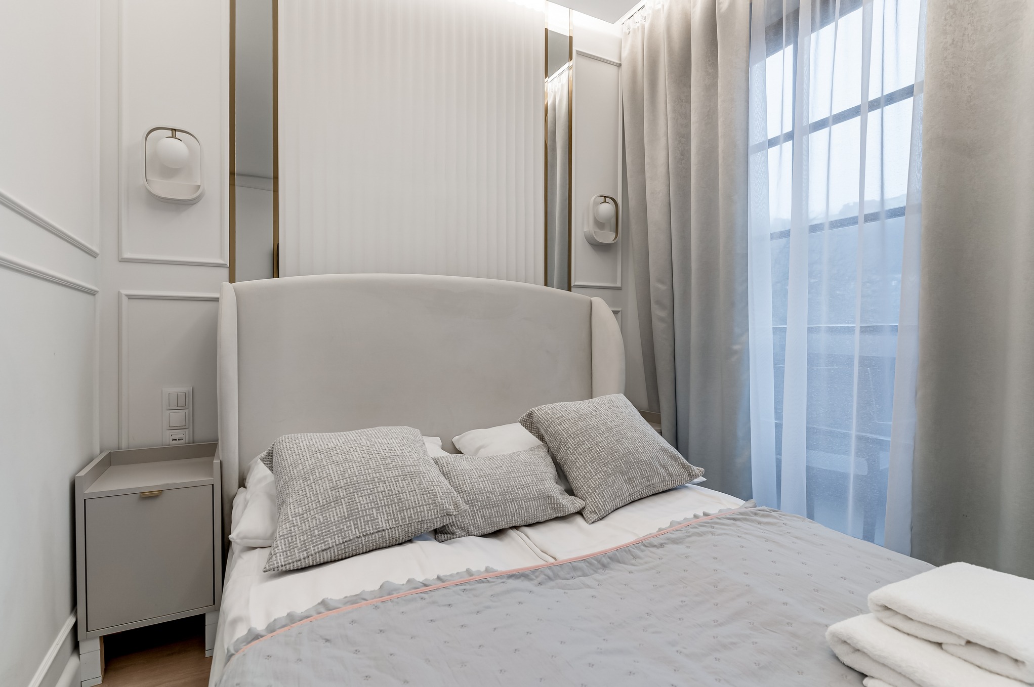 Jasna sypialnia w stylu glamour: łóżko z wysokim zagłówkiem, szare poduszki, zasłony i lampki nocne. Eleganckie wykończenie ścian z subtelnym oświetleniem.
