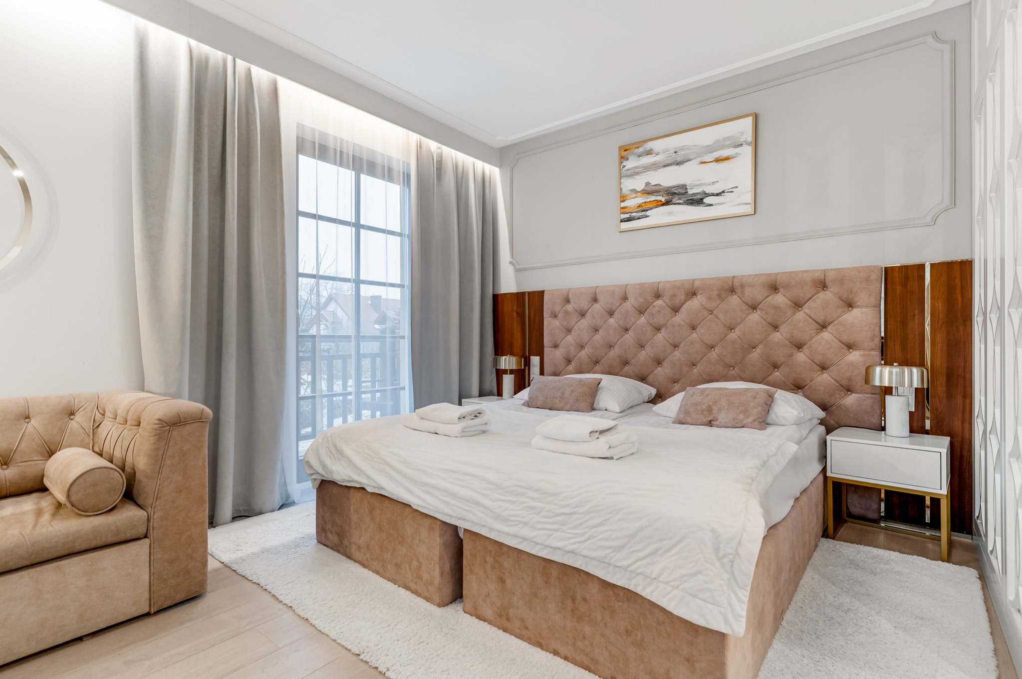 Elegancka sypialnia z pikowanym zagłówkiem, beżową sofą i dużym oknem z widokiem na zewnątrz. Na ścianie abstrakcyjny obraz w złotej ramie. Całość utrzymana w jasnej kolorystyce.