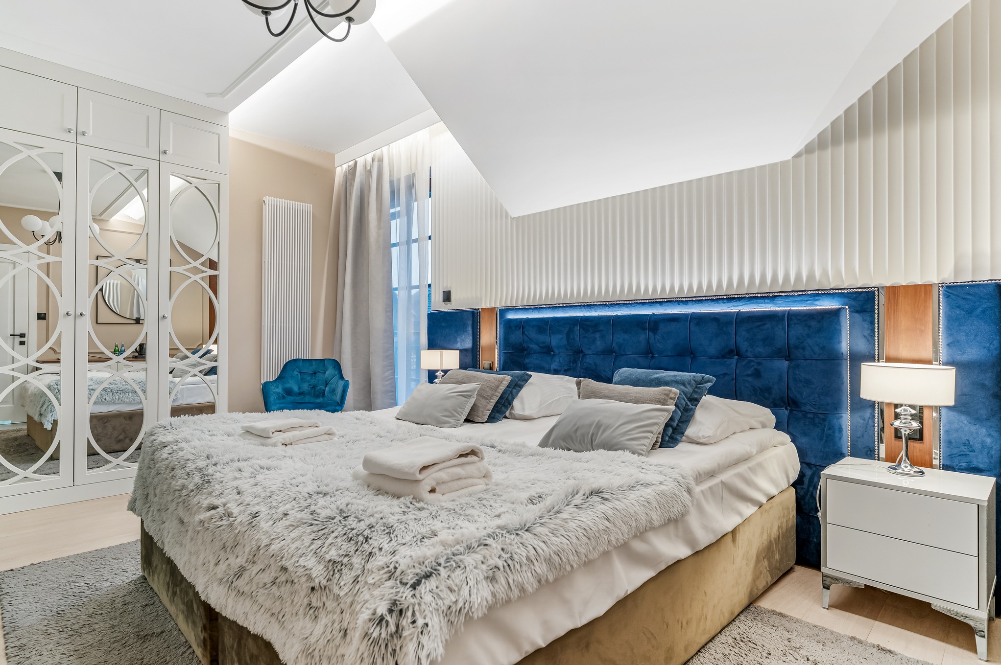 Elegancka sypialnia z łóżkiem o niebieskim, pikowanym wezgłowiu, puszystą narzutą i szafką nocną z lampką. W tle lustrzana szafa i krzesło w odcieniu turkusu.