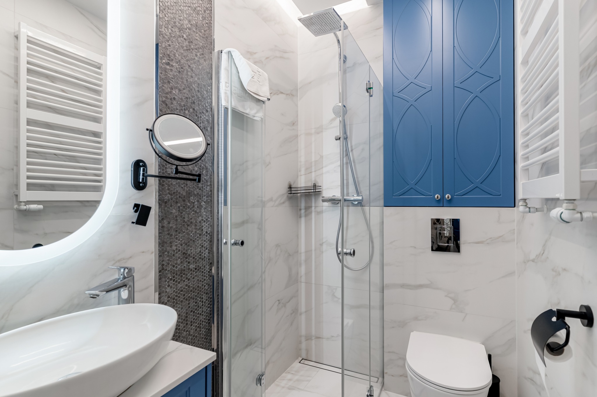 Nowoczesna łazienka z kabiną prysznicową typu walk-in, owalną umywalką na niebieskiej szafce i podświetlanym lustrem. Białe marmurowe płytki i niebieska szafka.