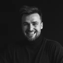 Portret uśmiechniętego mężczyzny z brodą, w czarnym golfie, na ciemnym tle, z widocznymi nierównościami uzębienia.