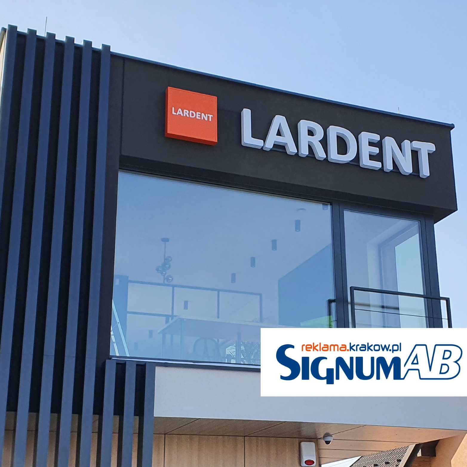 Zewnętrzny szyld firmy LARDENT z pomarańczowym kwadratem i białym napisem, widoczny z dołu, nowoczesna architektura budynku z czarnymi pionowymi listwami.