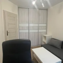 Jasne wnętrze pokoju z przesuwną szafą z płyty, z białymi wstawkami, sofą i stolikiem. Widoczna także drzwi i nowoczesna lampa sufitowa.