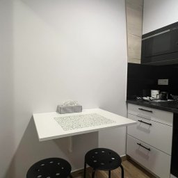 Bartosz Rząsa - Biała, składana półka kuchenna z czarnymi stołkami, na podłodze panele drewnopodobne. Ściana pomalowana na biało. Widok na część zabudowy kuchennej.