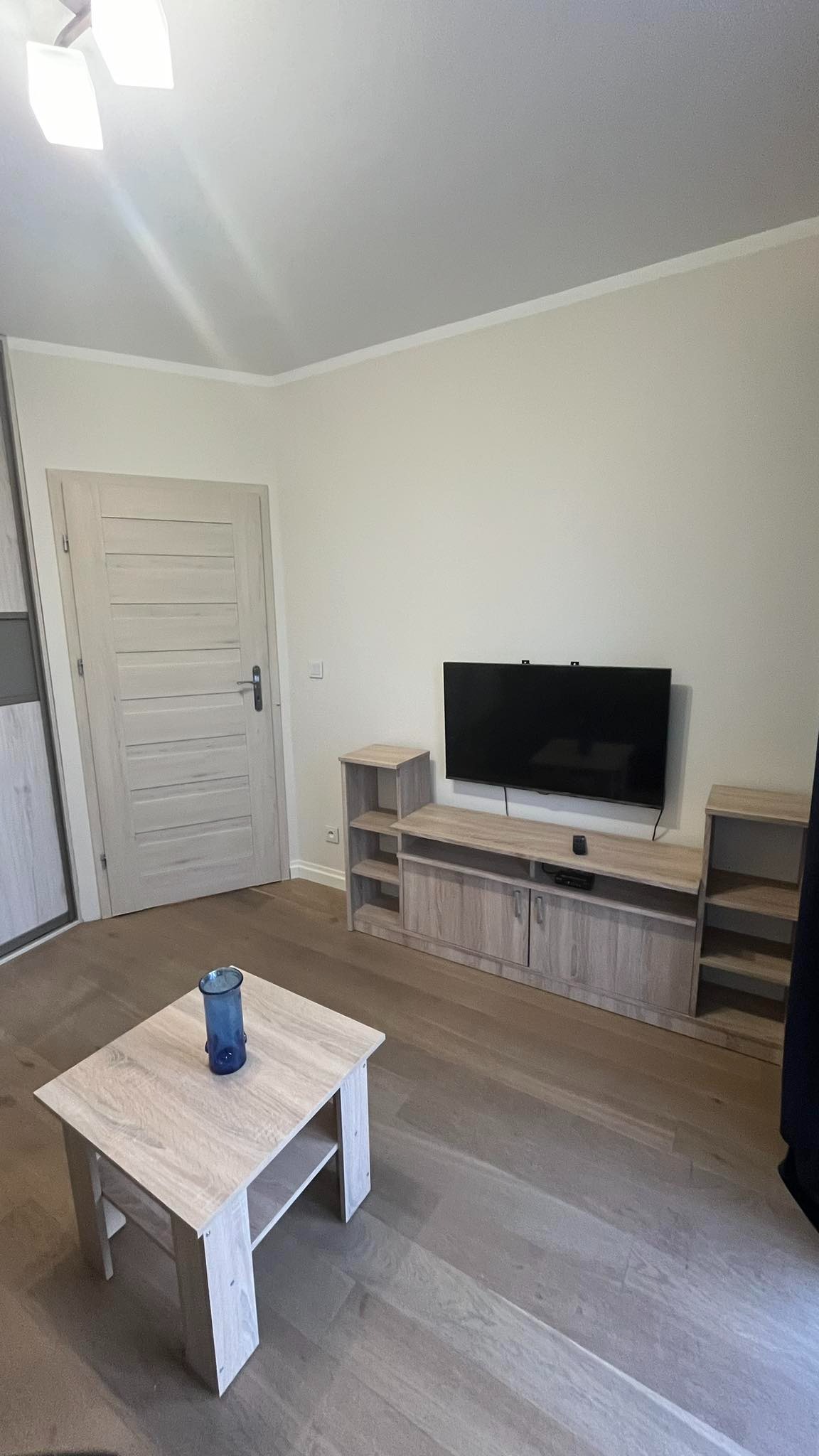 Jasne wnętrze pokoju po malowaniu: kremowe ściany, drewniana podłoga, meble w odcieniach beżu, telewizor na ścianie. Minimalistyczny styl.