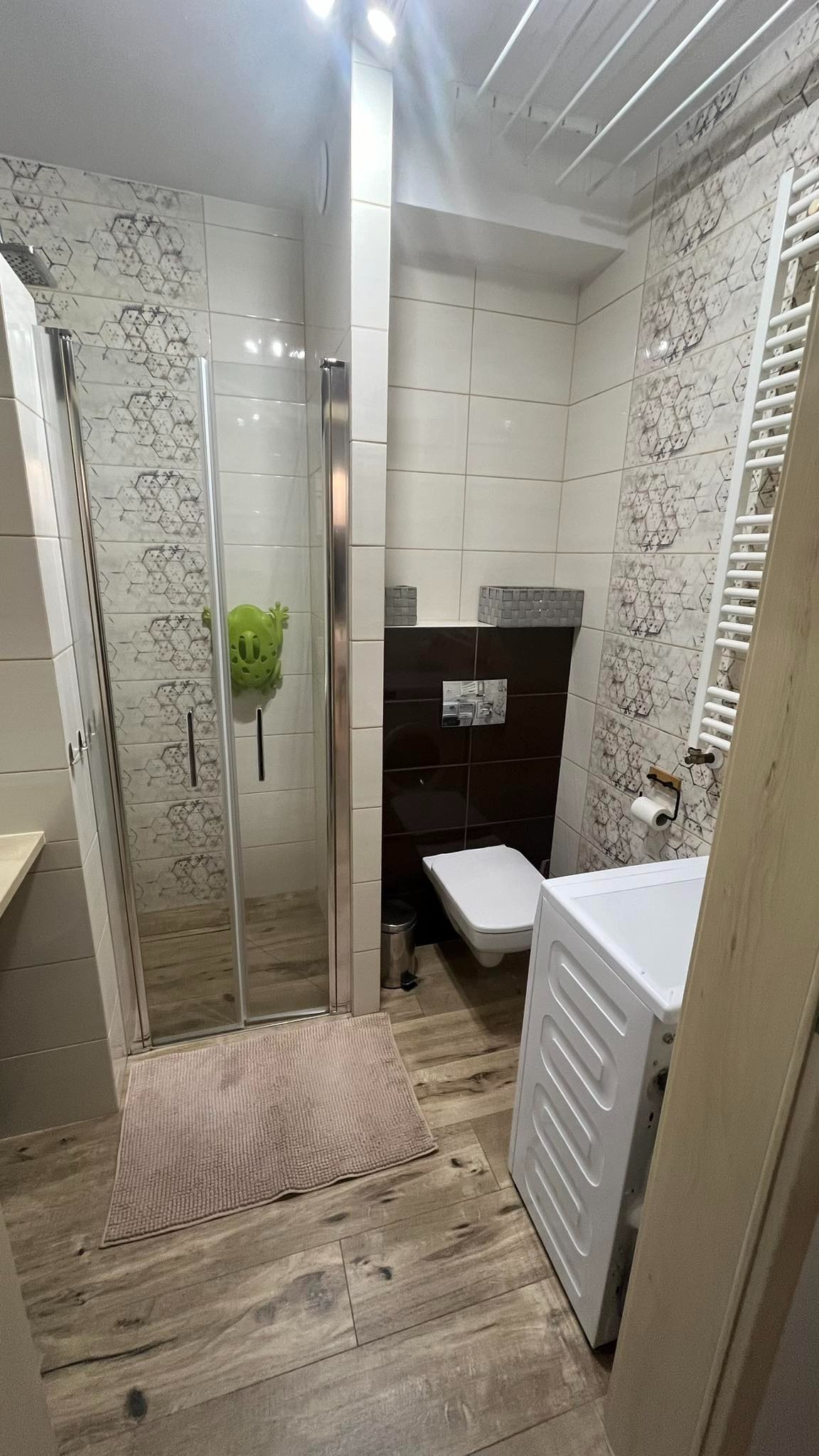 Ukończona łazienka z kabiną prysznicową, toaletą, pralką i grzejnikiem drabinkowym. Na ścianach białe i brązowe płytki. W kabinie prysznicowej zabawka w kształcie żaby.