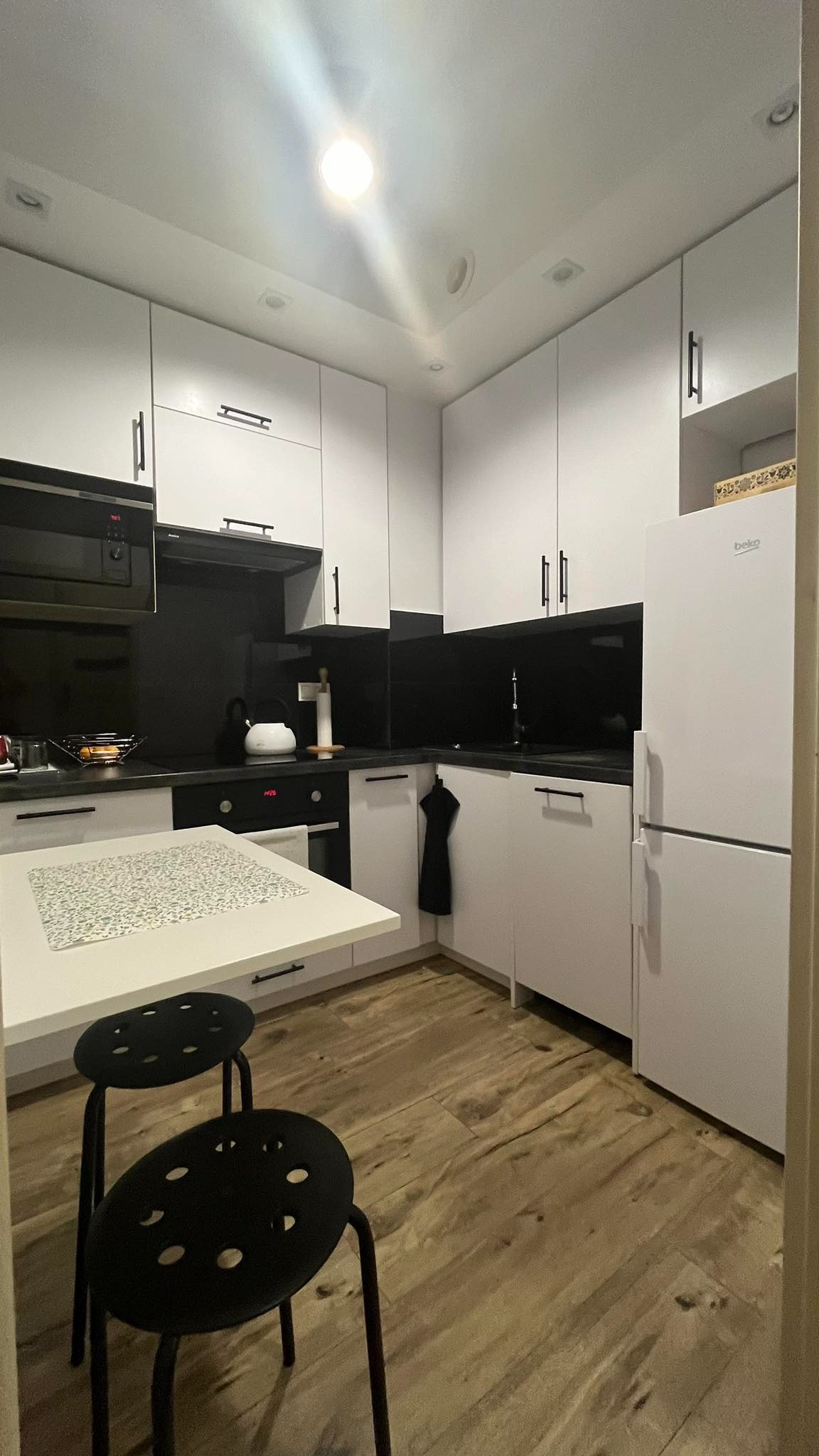 Nowoczesna, biała kuchnia z czarnym panelem nad blatem i drewnianą podłogą. Widoczne dwa czarne krzesła barowe i biały stół. Minimalistyczny design.