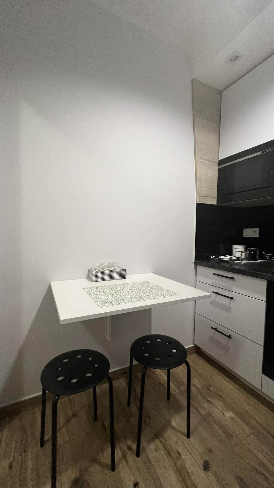 Biała, składana półka kuchenna z czarnymi stołkami, na podłodze panele drewnopodobne. Ściana pomalowana na biało. Widok na część zabudowy kuchennej.