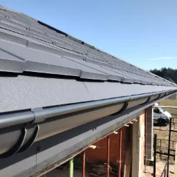 Nowe, grafitowe pokrycie dachowe z blachy trapezowej, widoczne rynny, dom w stanie surowym, rusztowania, błękitne niebo w tle.