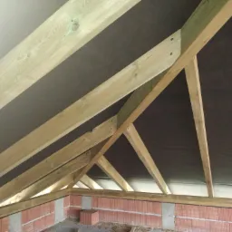 Surowa drewniana konstrukcja dachu z czarną membraną dachową widziana od wewnątrz budynku, z fragmentem murowanej ściany z czerwonej cegły.