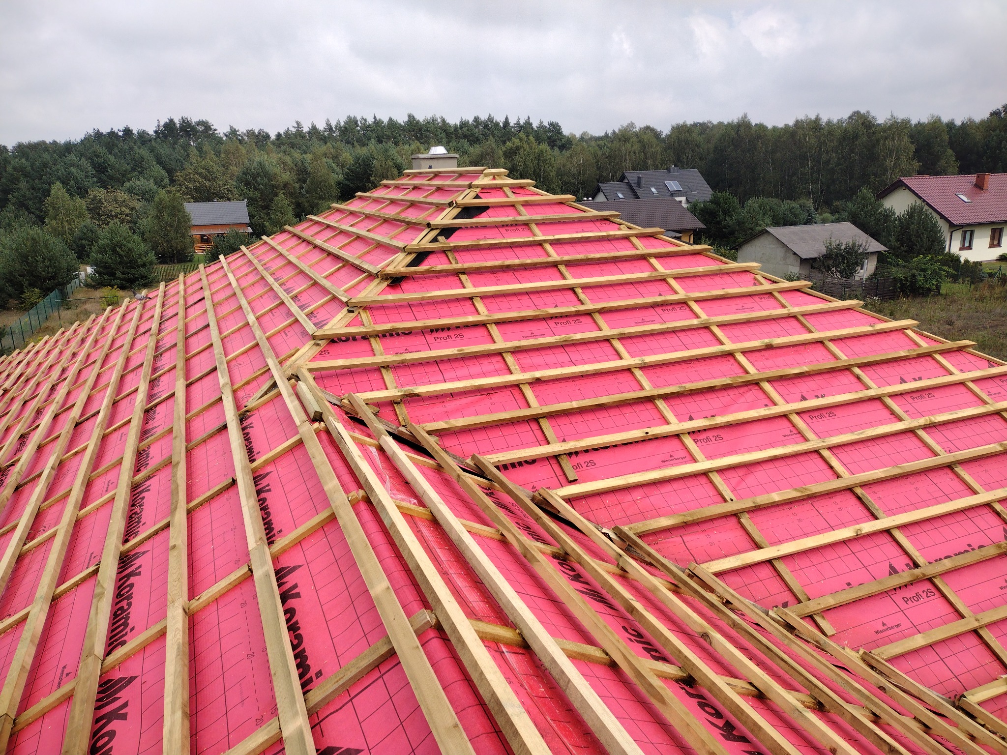 Wykonywanie konstrukcji drewnianej dachu z użyciem różowej membrany dachowej, widok z góry na tle drzew i domów.