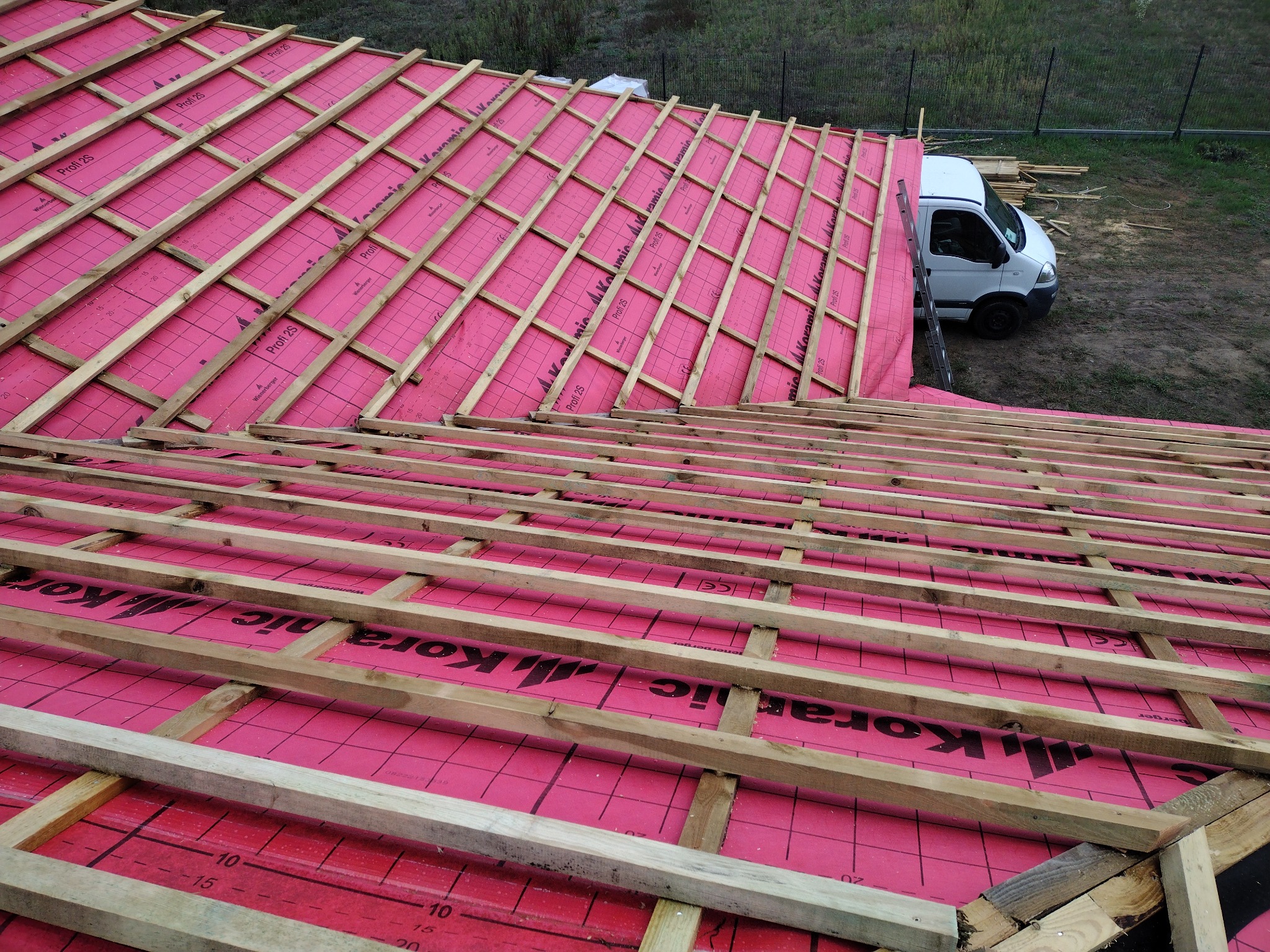 Widok z góry na konstrukcję dachu z drewnianą więźbą i różową membraną dachową, w tle zaparkowany biały samochód dostawczy.