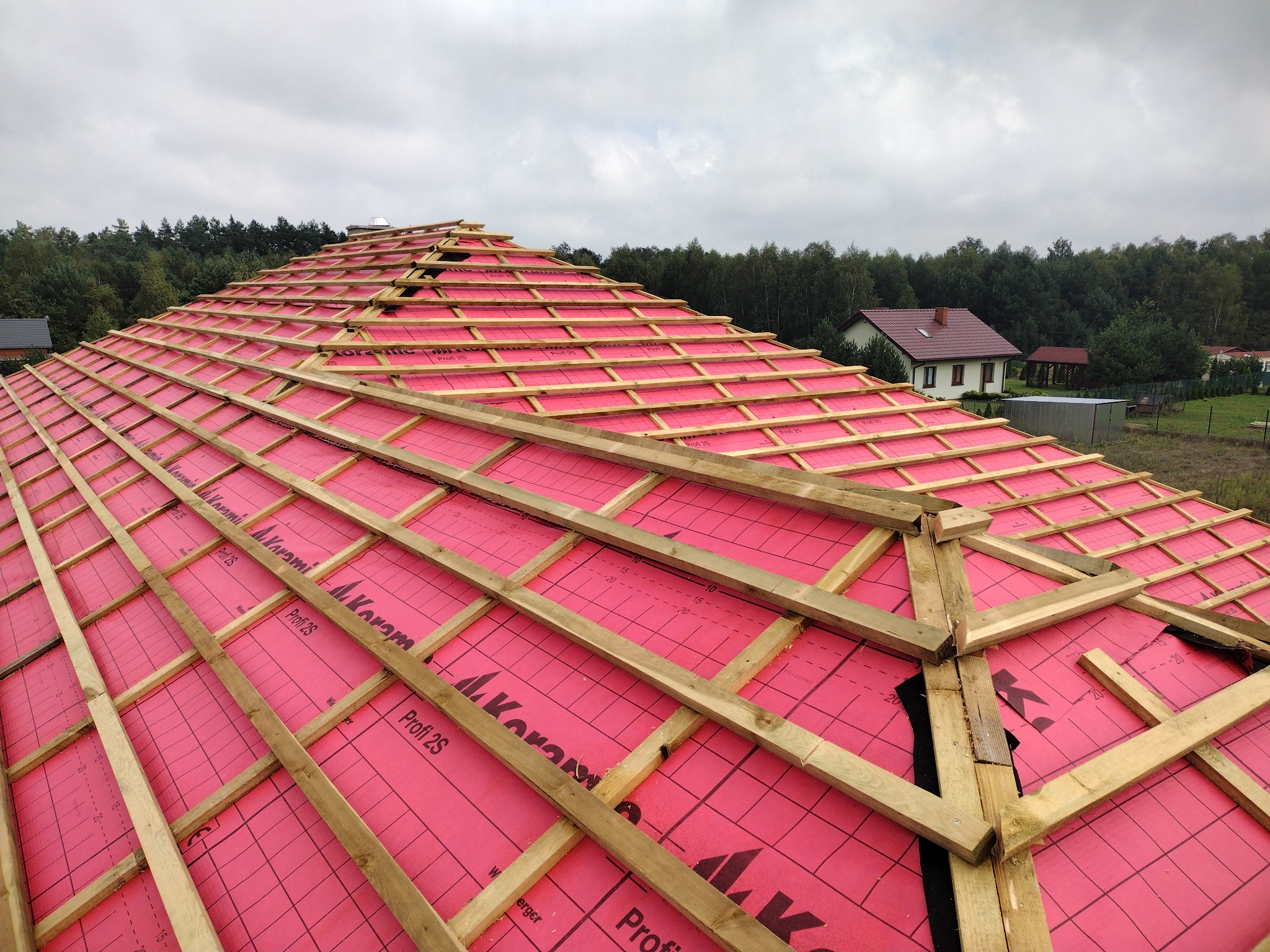 Więźba dachowa pokryta membraną w kolorze różowym z widocznymi oznaczeniami producenta, przygotowana pod krycie dachowe na tle zabudowy jednorodzinnej i lasu.