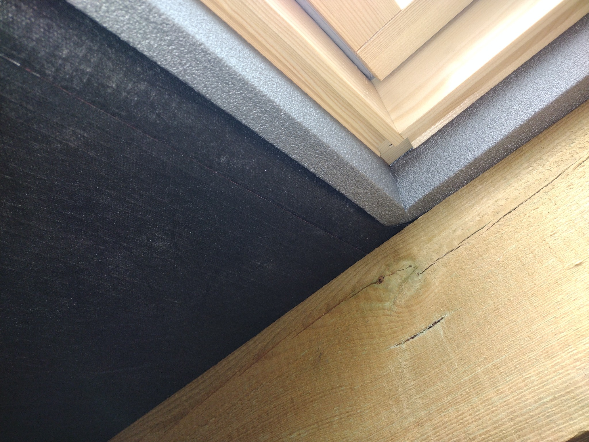 Szczegółowe ujęcie izolacji dachu z czarną membraną, szarą pianką uszczelniającą i drewnianą więźbą dachową z widocznym pęknięciem.