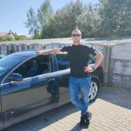 Mężczyzna w okularach przeciwsłonecznych opiera się o czarny samochód na tle betonowego ogrodzenia i drzew.