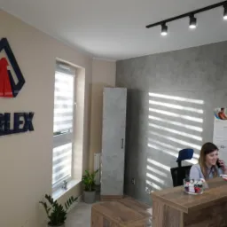 Recepcja firmy z logo KARLEX na ścianie, pracownica rozmawia przez telefon przy biurku, w tle szara ściana i rolety w oknie.