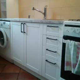 Biała zabudowa kuchenna z pralką pod blatem, czarne uchwyty, beżowe płytki na ścianie, widoczna część kuchenki z zawieszonymi ściereczkami.