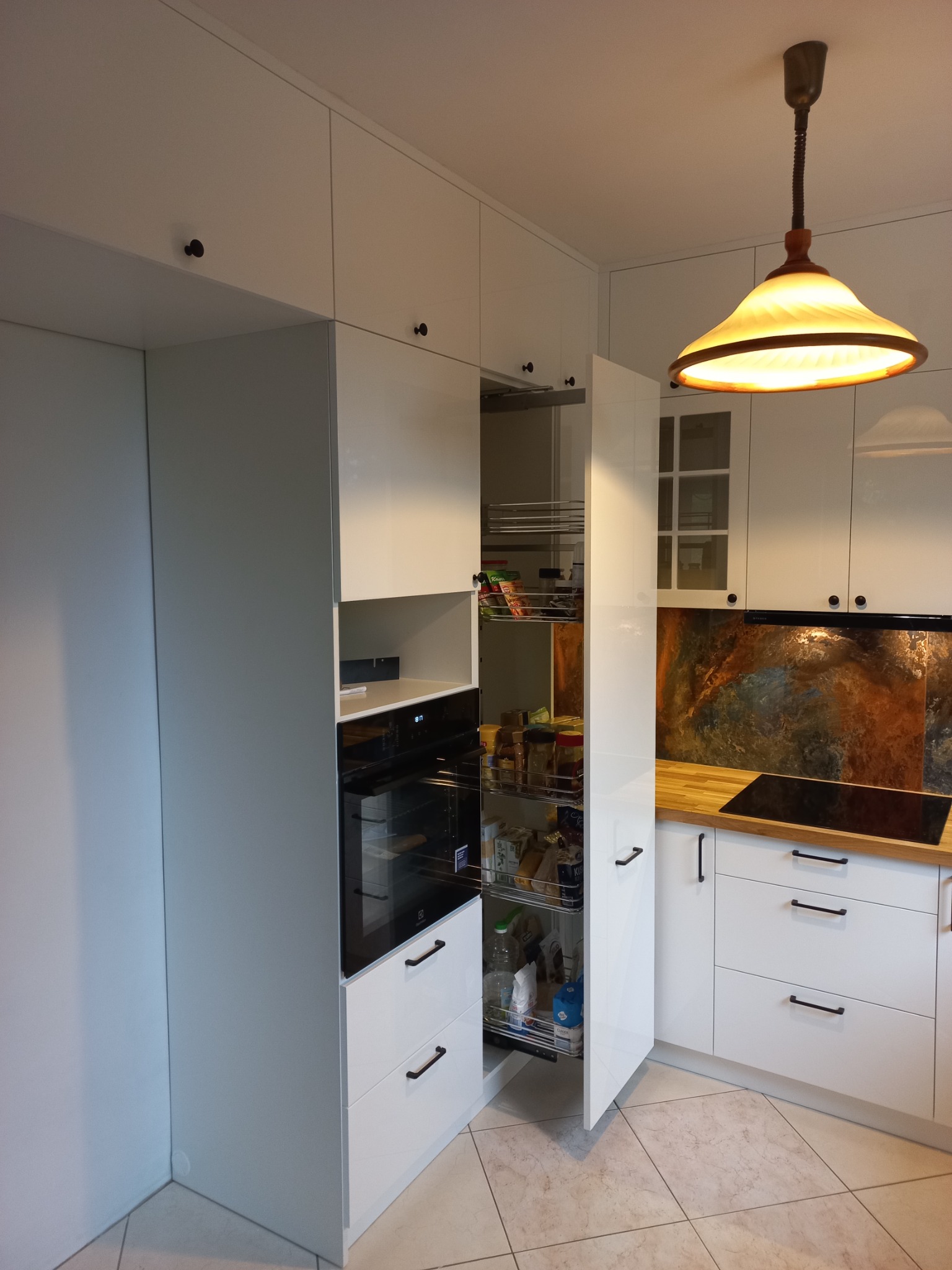 Biała, nowoczesna zabudowa kuchenna z wysuwaną spiżarnią, piekarnikiem i dekoracyjnym panelem nad blatem.