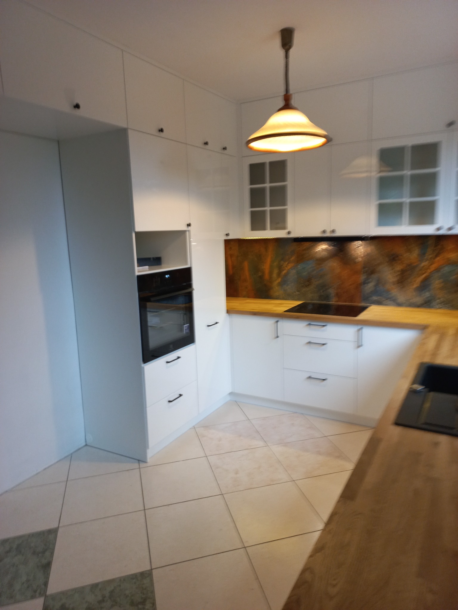 Nowoczesna, biała zabudowa kuchenna z drewnianym blatem i płytkami imitującymi kamień na ścianie w Mińsku Mazowieckim, oświetlona wiszącą lampą z abażurem w ciepłym odcieniu.