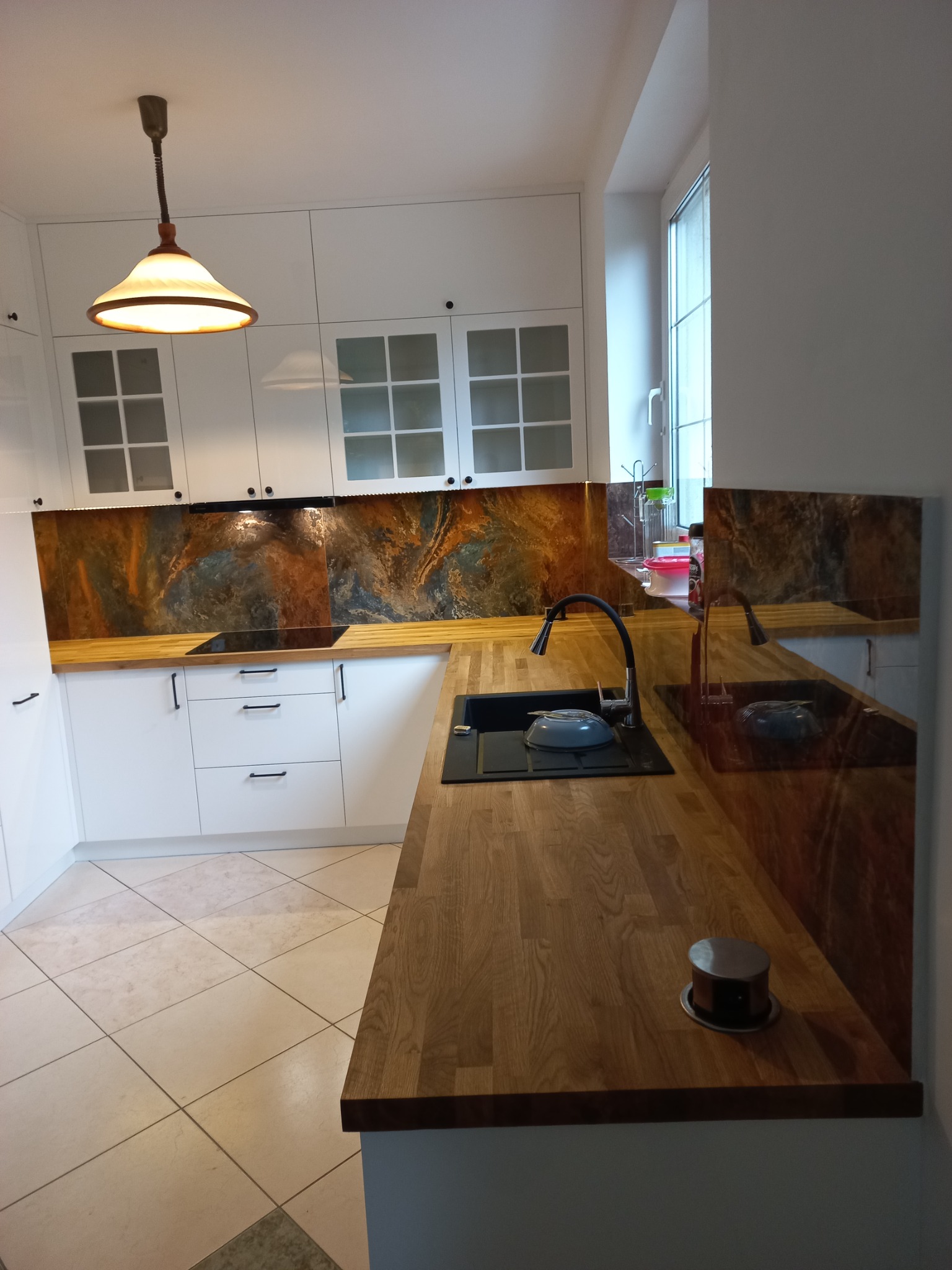 Nowoczesna kuchnia z białymi frontami szafek, drewnianym blatem i dekoracyjnym panelem ściennym imitującym kamień. Na blacie widoczny czarny zlew z baterią i szary talerz.