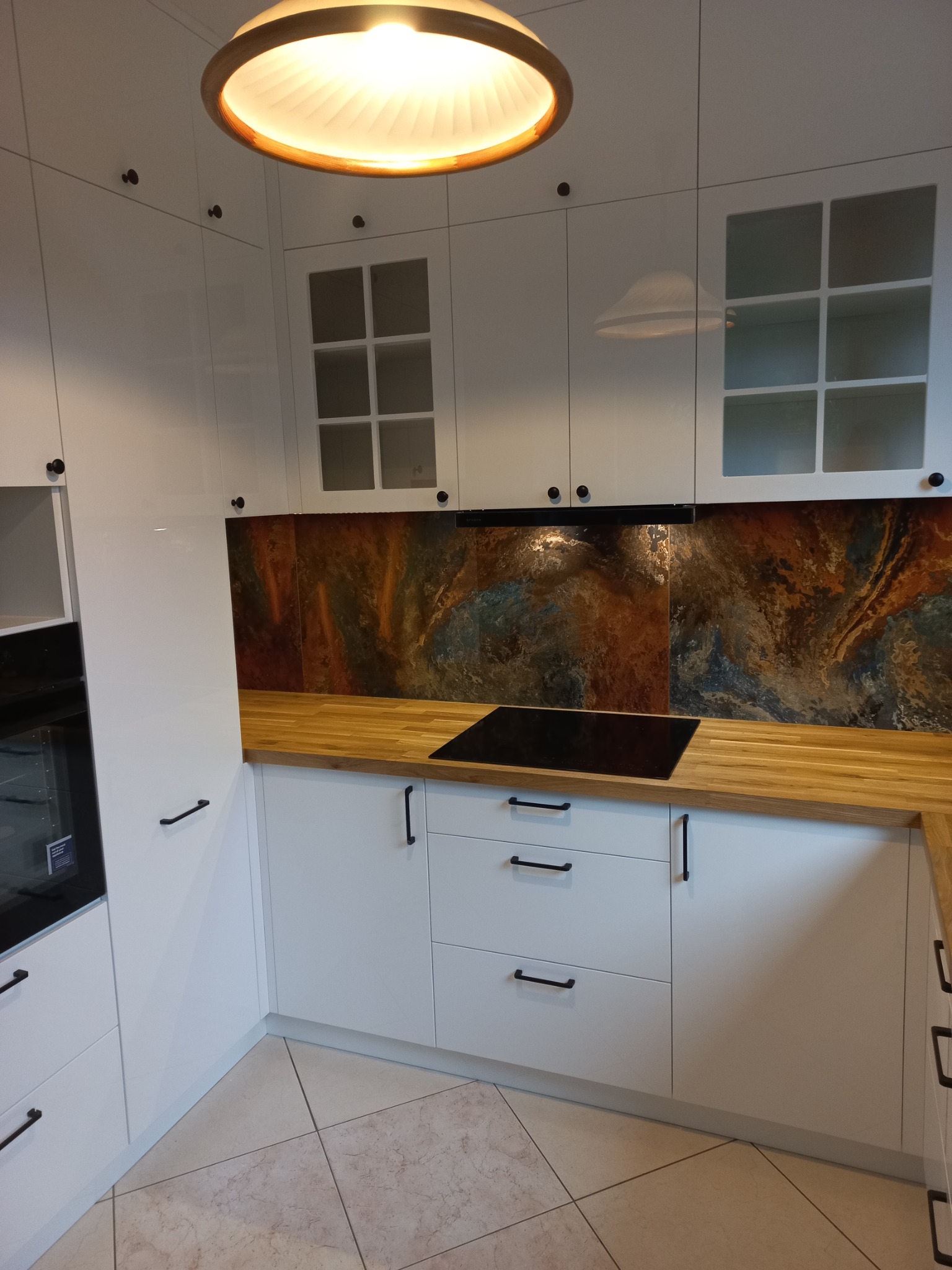 Biała kuchnia z drewnianym blatem i abstrakcyjnym panelem nad blatem, czarne uchwyty i okrągła lampa sufitowa