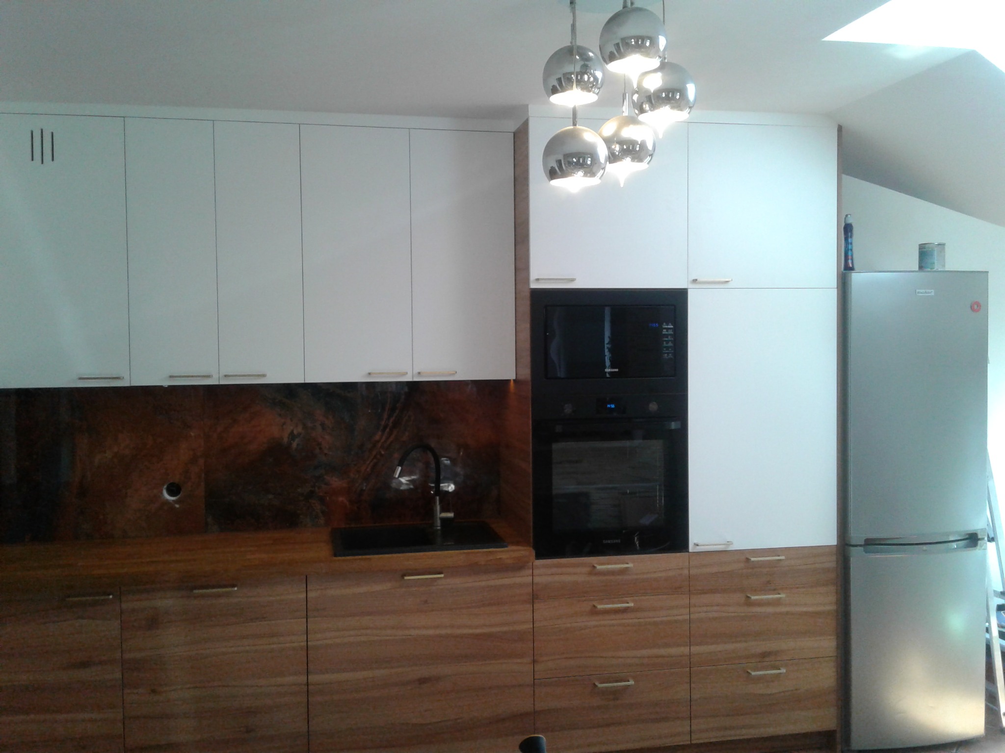 Nowoczesna kuchnia z białymi szafkami górnymi i drewnianymi dolnymi, zintegrowana z piekarnikiem i lodówką, oświetlona wiszącymi lampami w kształcie kul.