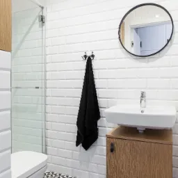 Nowoczesna łazienka z białą armaturą, ścianami wyłożonymi białymi, prostokątnymi płytkami w stylu metro i czarno-białą podłogą w geometryczne wzory. Na ścianie okrągłe lustro z czarną ramą i czarny...