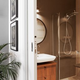 StudioRR MONIKA ROGOZIK-ROMANOWSKA - Nowoczesna łazienka z kabiną prysznicową, umywalką na drewnianej szafce i okrągłym lustrem, doświetlona punktowym oświetleniem, uzupełniona o roślinę doniczkową i grafiki na ścianie, Gdańsk.