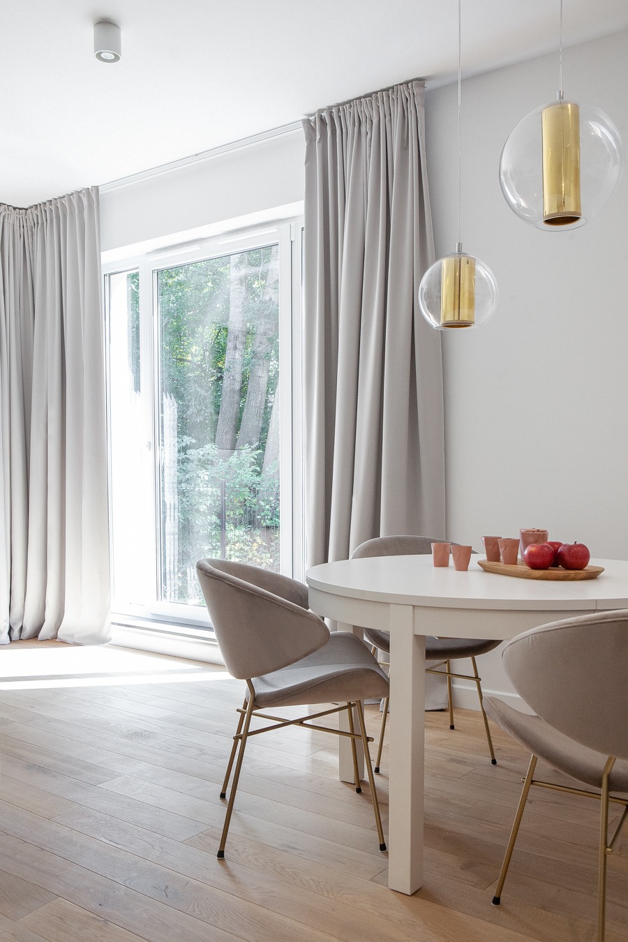 Jasne wnętrze jadalni z okrągłym białym stołem, welurowymi krzesłami na złotych nóżkach, drewnianą podłogą i szarymi zasłonami, oświetlone wiszącymi lampami z elementami w kolorze złota.
