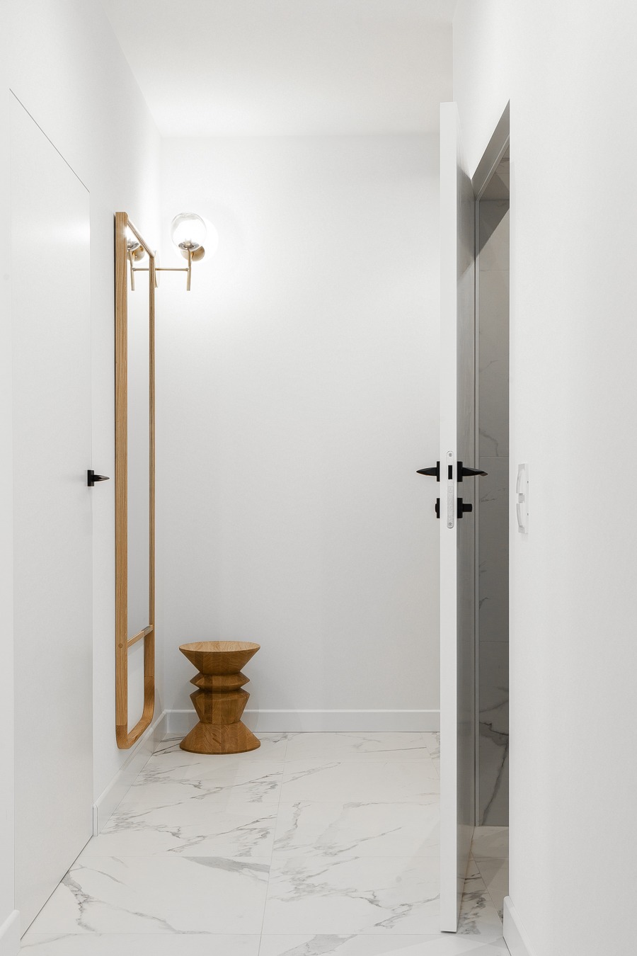 Minimalistyczny przedpokój w bieli z marmurową podłogą, drewnianym stołkiem klepsydrą i lustrem z oświetleniem kulistym. Otwarta biała rama drzwi ukazuje fragment łazienki z szarymi płytkami.