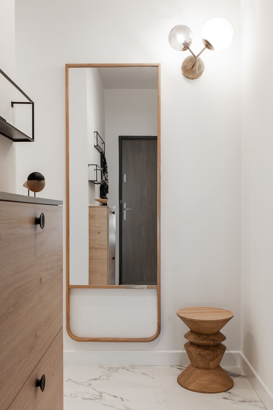 Nowoczesny przedpokój z drewnianymi meblami: szafka na buty z dekoracyjną figurką ptaka, lustro w drewnianej ramie odbijające drzwi wejściowe i drewniany stołek w kształcie klepsydry na marmurowej...