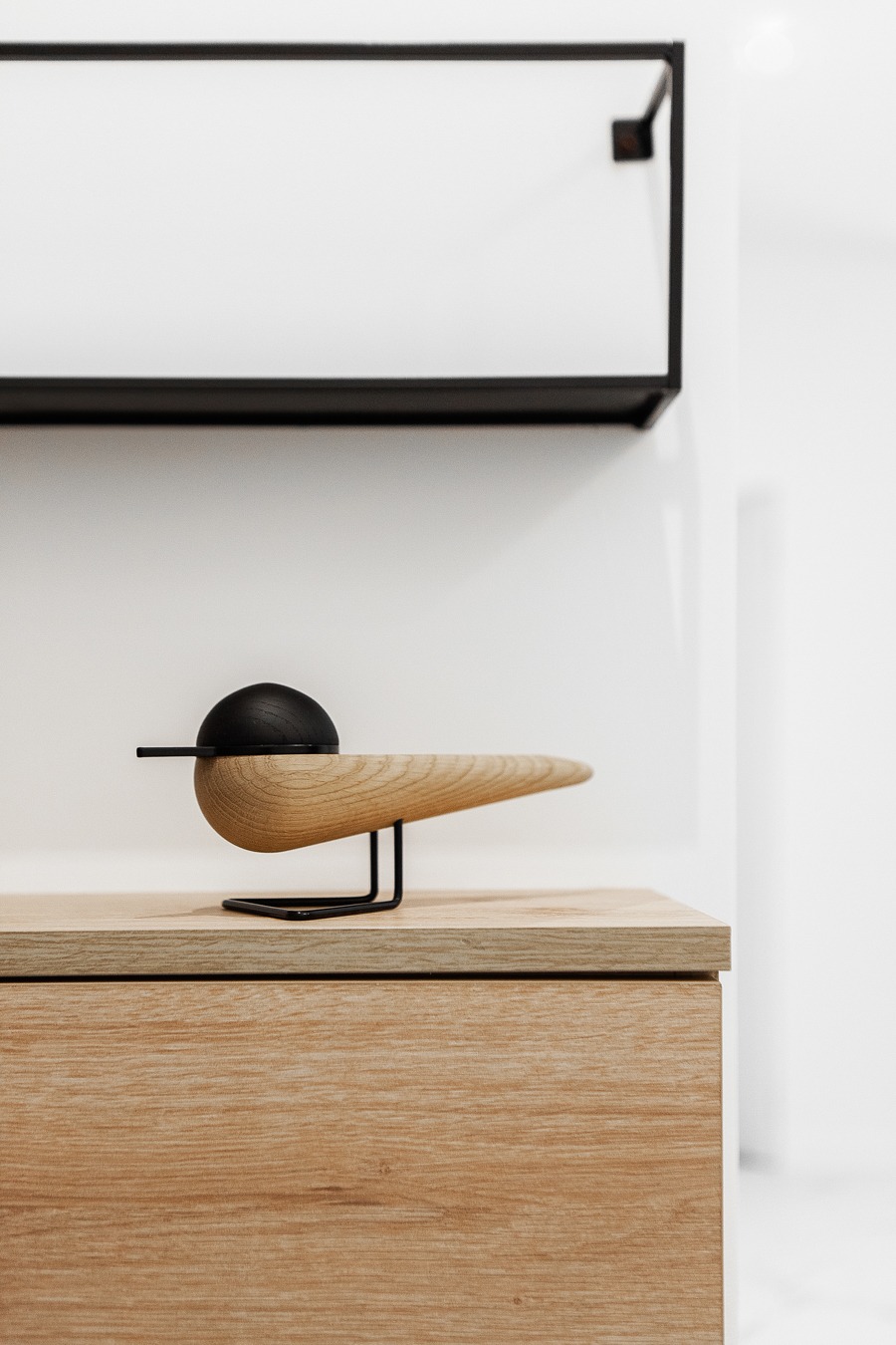 Drewniana figurka ptaka z czarnymi elementami na komodzie z jasnego drewna, z czarną półką na białej ścianie w tle.