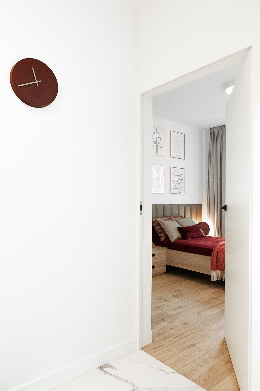 Widok na sypialnię przez otwarte drzwi: drewniana podłoga, łóżko z bordową pościelą, szara zasłona i grafiki na ścianie, brązowy zegar na białej ścianie po lewej.