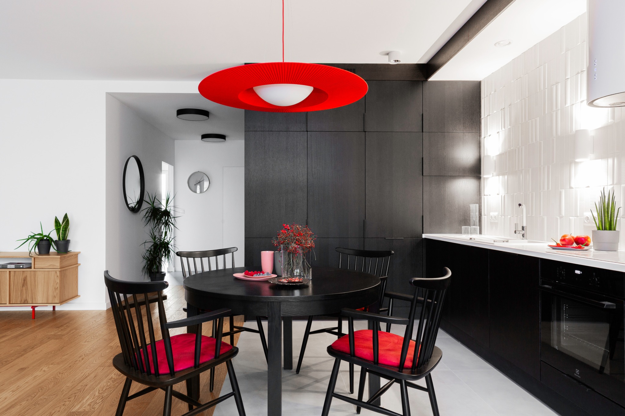 Nowoczesna jadalnia z czarnym okrągłym stołem, krzesłami z czerwonymi poduszkami i dużą czerwoną lampą wiszącą, połączona z kuchnią z czarnymi szafkami i białymi płytkami na ścianie, widok na salon...