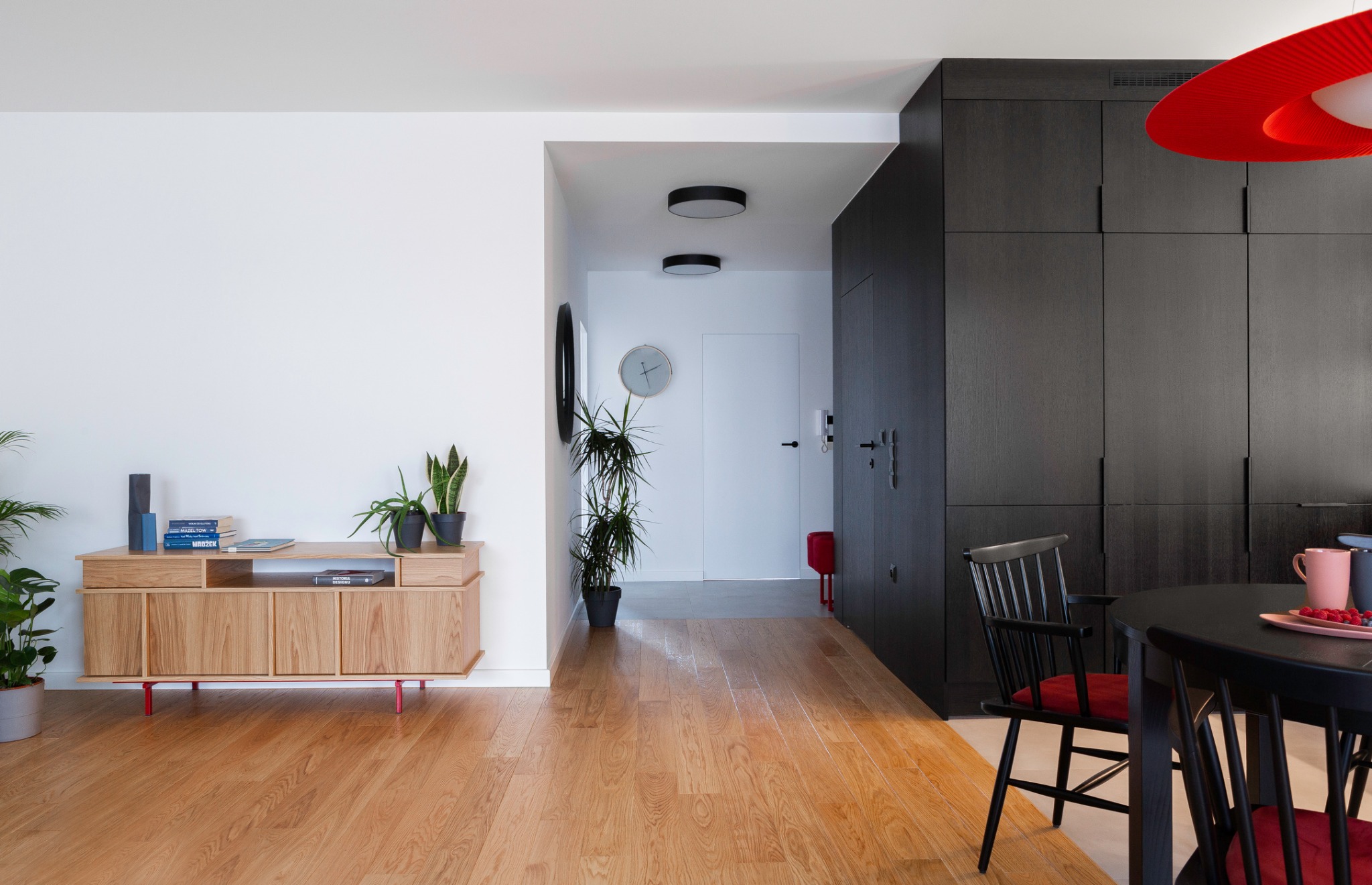 Jasne drewniane podłogi w mieszkaniu na Żoliborzu, komoda z książkami i roślinami, czarne szafy w zabudowie, okrągły stół z krzesłami i czerwonymi poduszkami, czerwona lampa wisząca, widok...