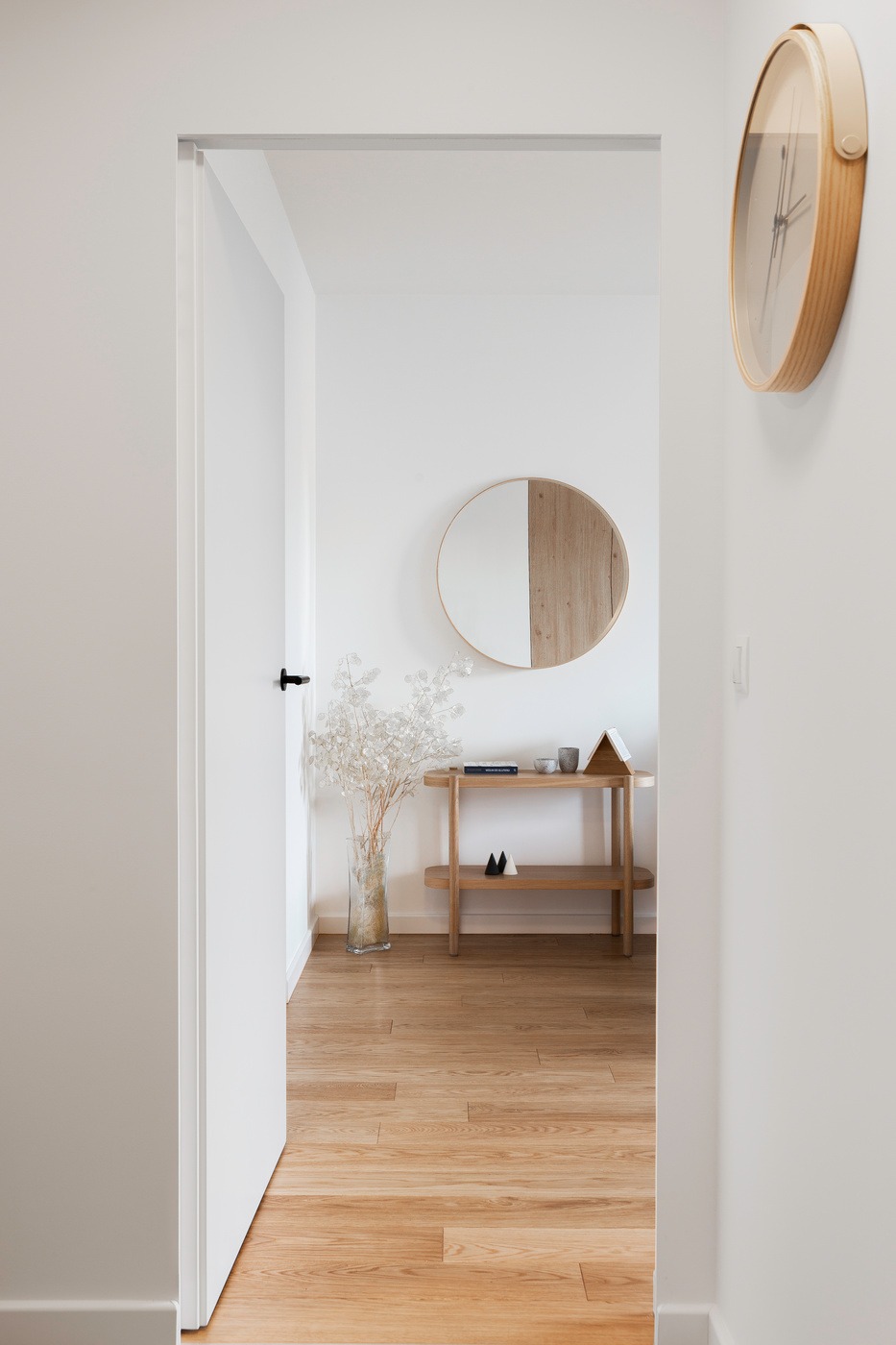 Nowoczesny przedpokój w mieszkaniu na Żoliborzu: drewniana podłoga, białe ściany, okrągłe lustro z podziałem na drewno i biel, drewniana konsola z dekoracjami i szklany wazon z suchymi gałązkami...