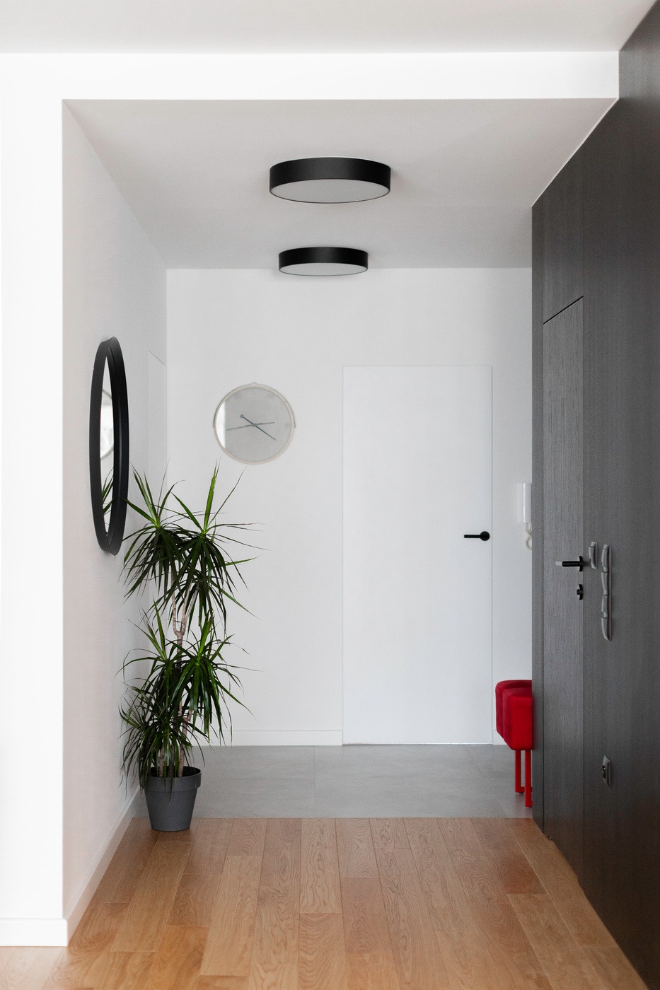 Nowoczesny przedpokój: drewniana podłoga przechodzi w szary gres, białe ściany z okrągłym lustrem i zegarem, doniczkowa roślina, czarne okrągłe lampy sufitowe, białe drzwi i fragment ciemnej szafy...